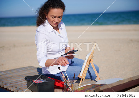 Beautiful woman sitting on a chaise lounge on the beach with smartphone in her hand. Painting tools on the foreground. Beautiful woman sitting on a chaise lounge on the beach with smartphone in her hand. Painting tools on the foreground. 78648692