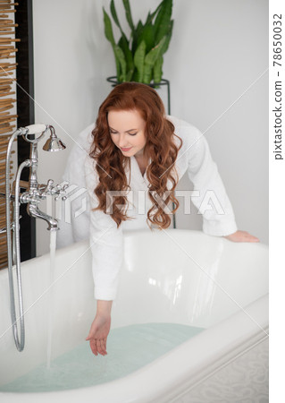 Pretty ginger woman in a white bathrobe getting a bath ready Pretty ginger woman in a white bathrobe getting a bath ready 78650032