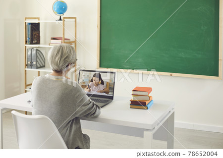 Online kids education. A female teacher with gray hair teaches a little schoolgirl has a laptop video call in class. Online kids education. A female teacher with gray hair teaches a little schoolgirl has a laptop video call in class. 78652004