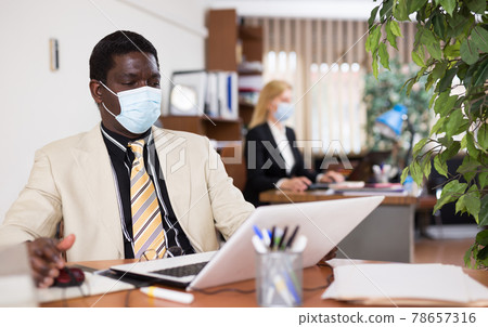 Focused african american businessman in a protective mask works at a computer in the office Focused african american businessman in a protective mask works at a computer in the office 78657316