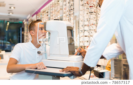 Teenage boy receiving eye examination on modern refractometer in optical store with professional African-American ophthalmologist Teenage boy receiving eye examination on modern refractometer in optical store with professional African-American ophthalmologist 78657317