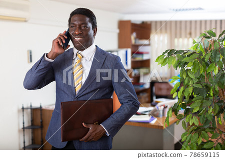 Smiling businessman with portfolio in hands talking on phone in office 78659115