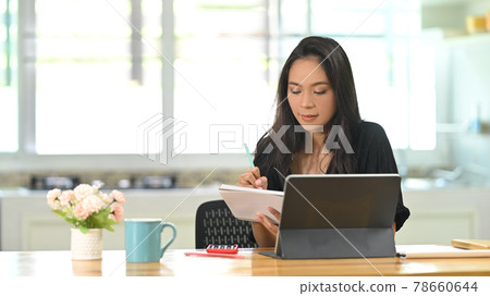 Young female freelancer taking note and working with tablet at home office. Young female freelancer taking note and working with tablet at home office. 78660644