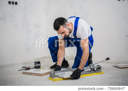 A worker in a blue overalls cuts a tile with a cutter A worker in a blue overalls cuts a tile with a cutter 78660647