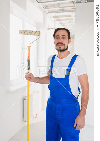 A bearded man in a blue overalls holds a roller in his hand and poses at the camera 78660681