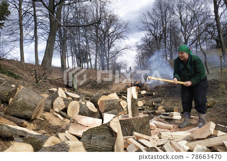 An old woman of a tavern shakes a firewood with an ax in the yard, prepares them for the winter. An old woman of a tavern shakes a firewood with an ax in the yard, prepares them for the winter. 78663470
