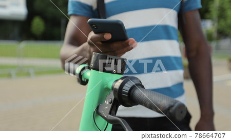 Young afro american man rents electric scooter using mobile phone app. The Man unlocked the electric scooter with his smartphone. Close-up. Young afro american man rents electric scooter using mobile phone app. The Man unlocked the electric scooter with his smartphone. Close-up. 78664707