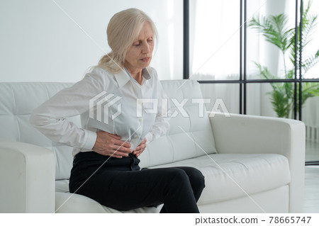 An elderly woman suffers from abdominal pain, a mature elderly adult woman feels abdominal pain while sitting on the couch An elderly woman suffers from abdominal pain, a mature elderly adult woman feels abdominal pain while sitting on the couch 78665747