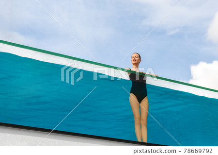 Young adult caucasian sporty slim smiling relaxing woman enjoy stand in pool with clear blue water and trasparent wall edge against sky at luxury spa resort. Healthy lifestyle and wellness concept Young adult caucasian sporty slim smiling relaxing woman enjoy stand in pool with clear blue water and trasparent wall edge against sky at luxury spa resort. Healthy lifestyle and wellness concept 78669792