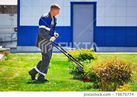 A young man walks with a lawn mower on a green lawn in a production area. A young man walks with a lawn mower on a green lawn in a production area. 78670774