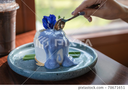 Woman hand using spoon cutting coconut cake with butterfly pea topping on ceramic dish and iced cocoa on the table 78671651