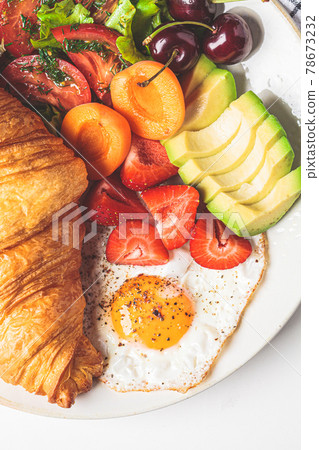 Breakfast plate with croissant, avocado, fried egg, salad and fruit. 78673232