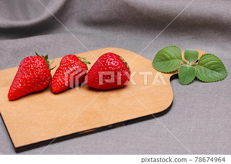 Three ripe red delicious strawberries lie in a row on a wooden cutting board next to a mint leaf. Summer harvest. Summer time. 78674964