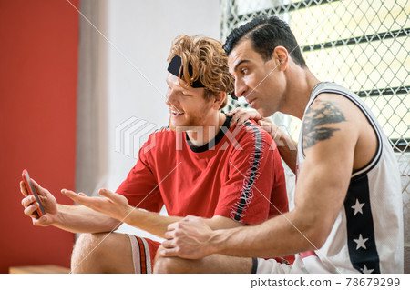 Two basketball players watching something on the smartphone and looking involved Two basketball players watching something on the smartphone and looking involved 78679299
