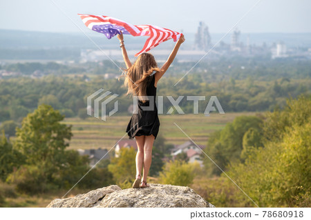 Young pretty american woman with long hair holding waving on wind USA flag on her sholders standing outdoors enjoying warm summer day. Young pretty american woman with long hair holding waving on wind USA flag on her sholders standing outdoors enjoying warm summer day. 78680918