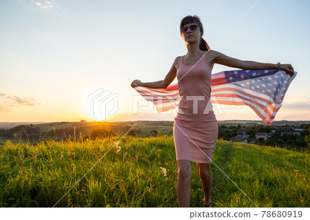Happy young woman posing with USA national flag standing outdoors at sunset. Positive female celebrating United States independence day. International day of democracy concept. Happy young woman posing with USA national flag standing outdoors at sunset. Positive female celebrating United States independence day. International day of democracy concept. 78680919