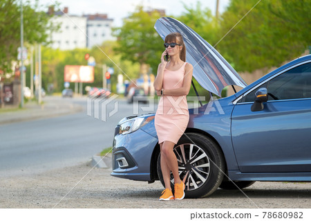 Helpless woman standing near her car with open hood takling on mobile phone calling road service for help. Young female driver having trouble with vehicle. Helpless woman standing near her car with open hood takling on mobile phone calling road service for help. Young female driver having trouble with vehicle. 78680982