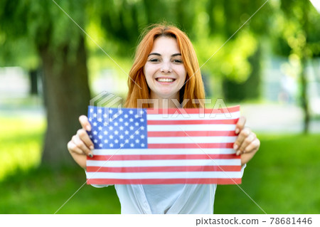 Portrait of happy young red haired woman holding USA national flag in her hands standing outdoors in summer park. Positive girl celebrating United States independence day. International day of Portrait of happy young red haired woman holding USA national flag in her hands standing outdoors in summer park. Positive girl celebrating United States independence day. International day of 78681446