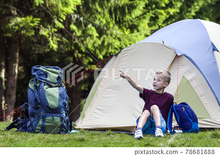 Holiday camping. Young boy sitting in front of a tent near backpacks taking rest after hiking in the forest shows something in the trees. Traveling and outdoors activities. Holiday camping. Young boy sitting in front of a tent near backpacks taking rest after hiking in the forest shows something in the trees. Traveling and outdoors activities. 78681888