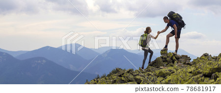 Young tourists with backpacks, athletic man helps slim woman to clime rocky mountain top against bright summer sky and mountain range background. Tourism, traveling and healthy lifestyle concept. 78681917