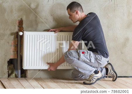 Young handsome professional plumber worker installing heating radiator in an empty room of a newly built apartment or house. Construction, maintenance and repair concept. Young handsome professional plumber worker installing heating radiator in an empty room of a newly built apartment or house. Construction, maintenance and repair concept. 78682001