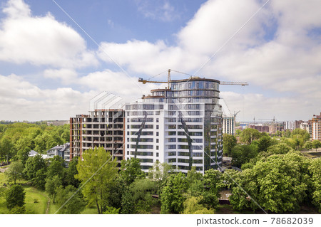 Apartment or office tall building unfinished under construction among green tree tops. Tower cranes on bright blue sky copy space background. Apartment or office tall building unfinished under construction among green tree tops. Tower cranes on bright blue sky copy space background. 78682039