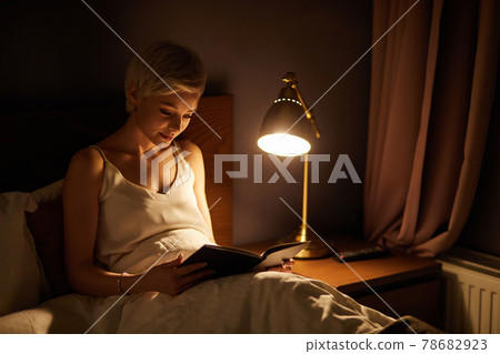 Beautiful female in pajamas lying on bed reading book, alone at night Beautiful female in pajamas lying on bed reading book, alone at night 78682923