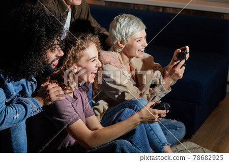 Happy excited friends having fun by playing game console at home, indoors. Friendship, leasure, rest, home party concept Happy excited friends having fun by playing game console at home, indoors. Friendship, leasure, rest, home party concept 78682925