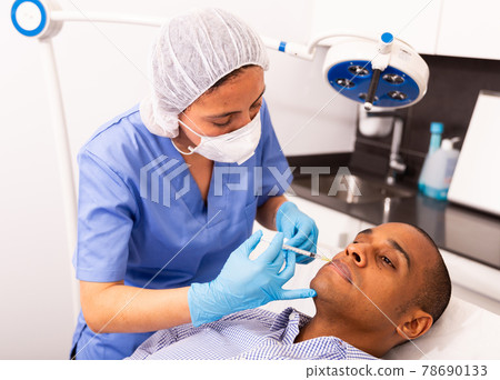 Female beautician injecting facial filler into skin for male client Female beautician injecting facial filler into skin for male client 78690133