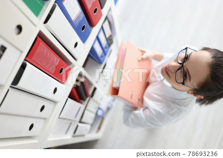 Young woman secretary in glasses takes folder with documents from shelf in archive 78693236