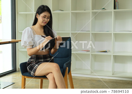 Smiling young female student with eyeglasses reading book in library 78694510