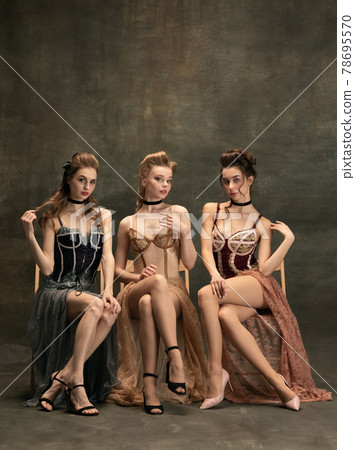 Pretty women in vintage 20s dresses. Young girls in art action sitting together and posing on wall background. 78695570