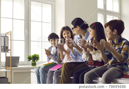 Female teacher together with group of happy children clapping hands while sitting in classroom. 78695686