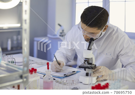 Scientist taking notes sitting at table with microscope in microbiology laboratory 78695750