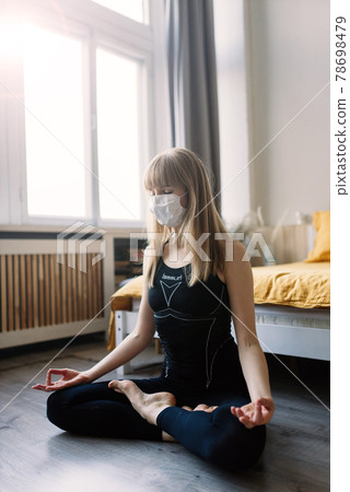 Meditating woman wearing a medical face mask to protect from corona virus, yoga position on mat home 78698479