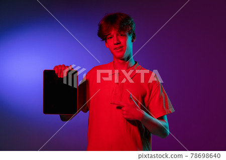 Holding tablet, crazy excited. Caucasian young man's portrait on black studio background in neon light. Beautiful male model in white shirt. Holding tablet, crazy excited. Caucasian young man's portrait on black studio background in neon light. Beautiful male model in white shirt. 78698640