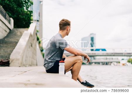 Tired Young man runner sitting and relaxing after sport training. Holding water bottle while doing workout in summer city street. 78699600