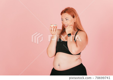 Oversized young woman wearing sports outfit looks at sweet donut junk food 78699741