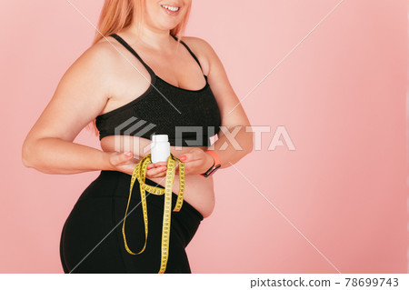 Happy young woman with oversized body wearing yoga outfit holding jar of pills 78699743