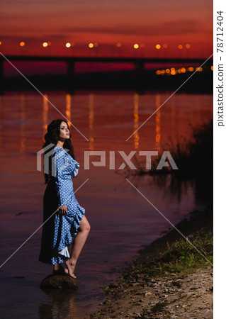 Beautiful young girl with long dark wavy hair standing at the bank of the river Beautiful young girl with long dark wavy hair standing at the bank of the river 78712404