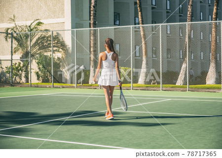 Tennis player woman playing on green hard court at resort hotel outside wearing white tennis dress outfit from behind. 78717365
