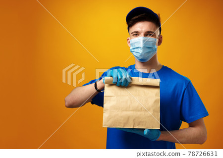 Young delivery man in face mask and gloves holding food delivery parcel against yellow background Young delivery man in face mask and gloves holding food delivery parcel against yellow background 78726631