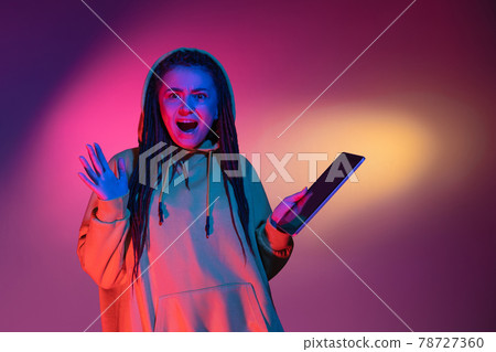 Holding tablet, crazy excited. Caucasian young woman's portrait on black studio background in neon light. Beautiful male model in white shirt. Holding tablet, crazy excited. Caucasian young woman's portrait on black studio background in neon light. Beautiful male model in white shirt. 78727360
