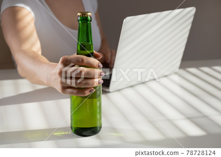 Caucasian woman works at a laptop and drinks beer from a glass bottle Caucasian woman works at a laptop and drinks beer from a glass bottle 78728124