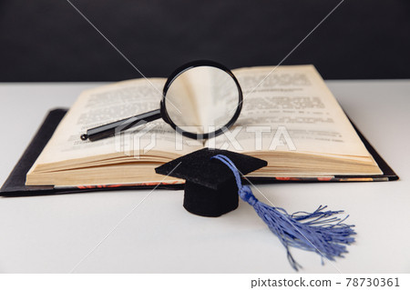 Magnifier on open book and graduation cap on white table. Education concept 78730361