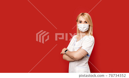 Confident female doctor wearing protective face mask standing on red background arms crossed. Young woman intern in medical clothes posing. 78734352