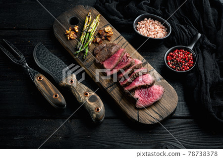 Grilled fillet mignon beef steak, with onion and asparagus, on wooden serving board, with meat knife and fork, on black wooden table background 78738378