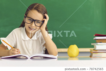 Happy elementary school girl student in eyeglasses sitting at desk and studying 78742565