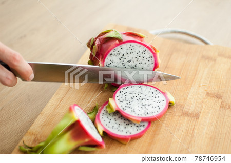 Female hands is cutting a dragon fruit or pitaya with pink skin and white pulp with black seeds on wooden cut board on the table. Exotic fruits, healthy eating concept Female hands is cutting a dragon fruit or pitaya with pink skin and white pulp with black seeds on wooden cut board on the table. Exotic fruits, healthy eating concept 78746954