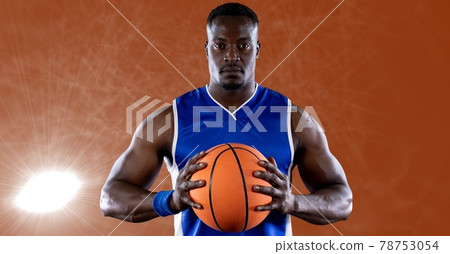 African american male basketball player holding basketball against spot of light in background African american male basketball player holding basketball against spot of light in background 78753054